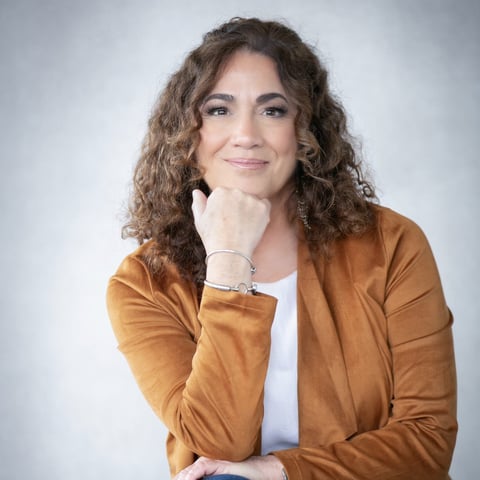 Woman with curly brown hair wearing a tan cardigan, hand to face, smiling at camera against white background